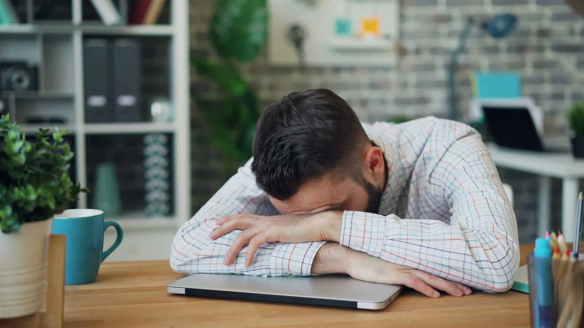 Man with his head resting on his arms at a desk beside a laptop, illustrating stress and uncertainty about Medicaid coverage, benefits, or eligibility.