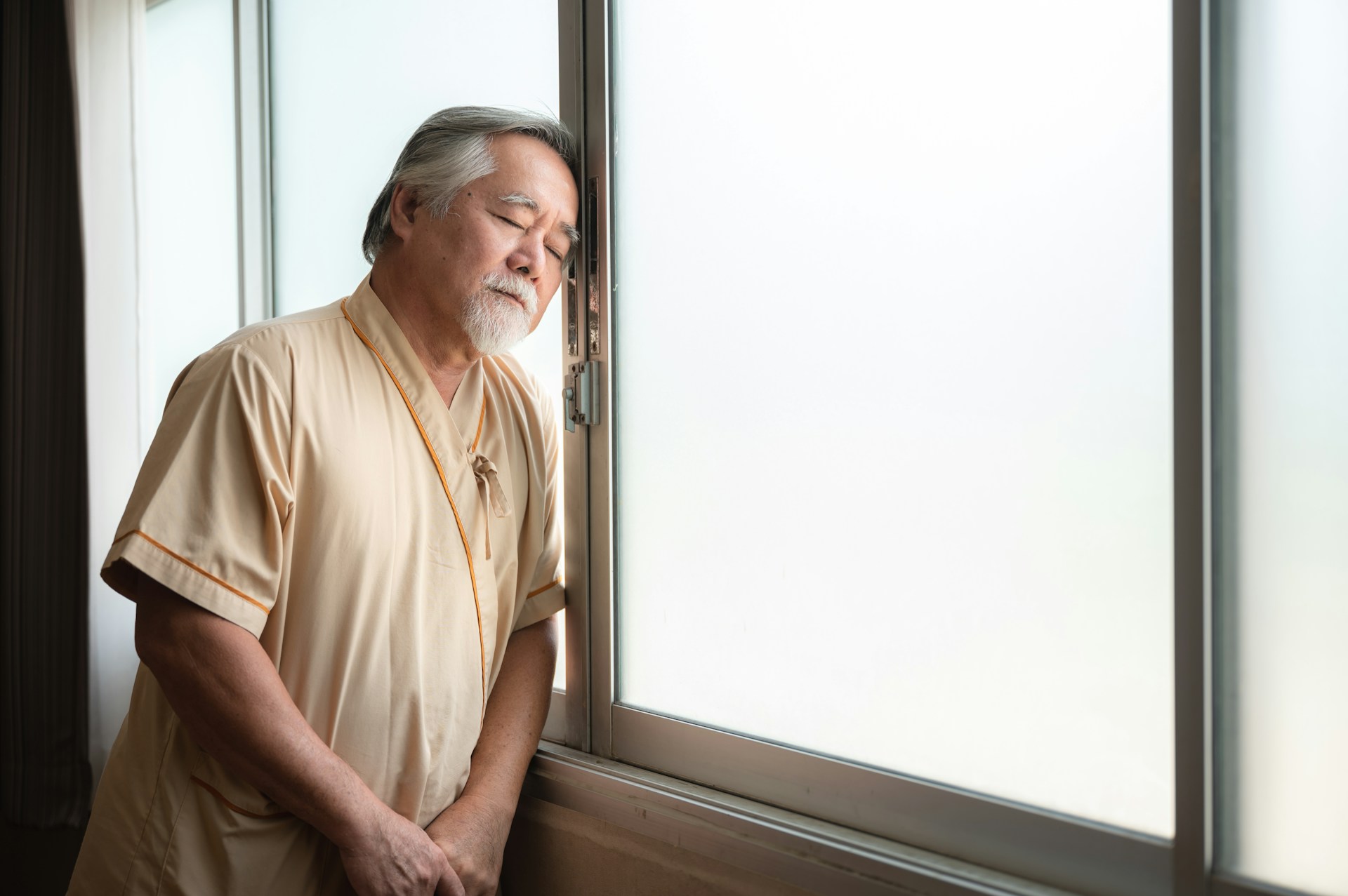 An elderly man with gray hair and a beard stands by a window with his eyes closed, appearing reflective and tired, suggesting themes of health and care in a quiet, intimate moment indoors.