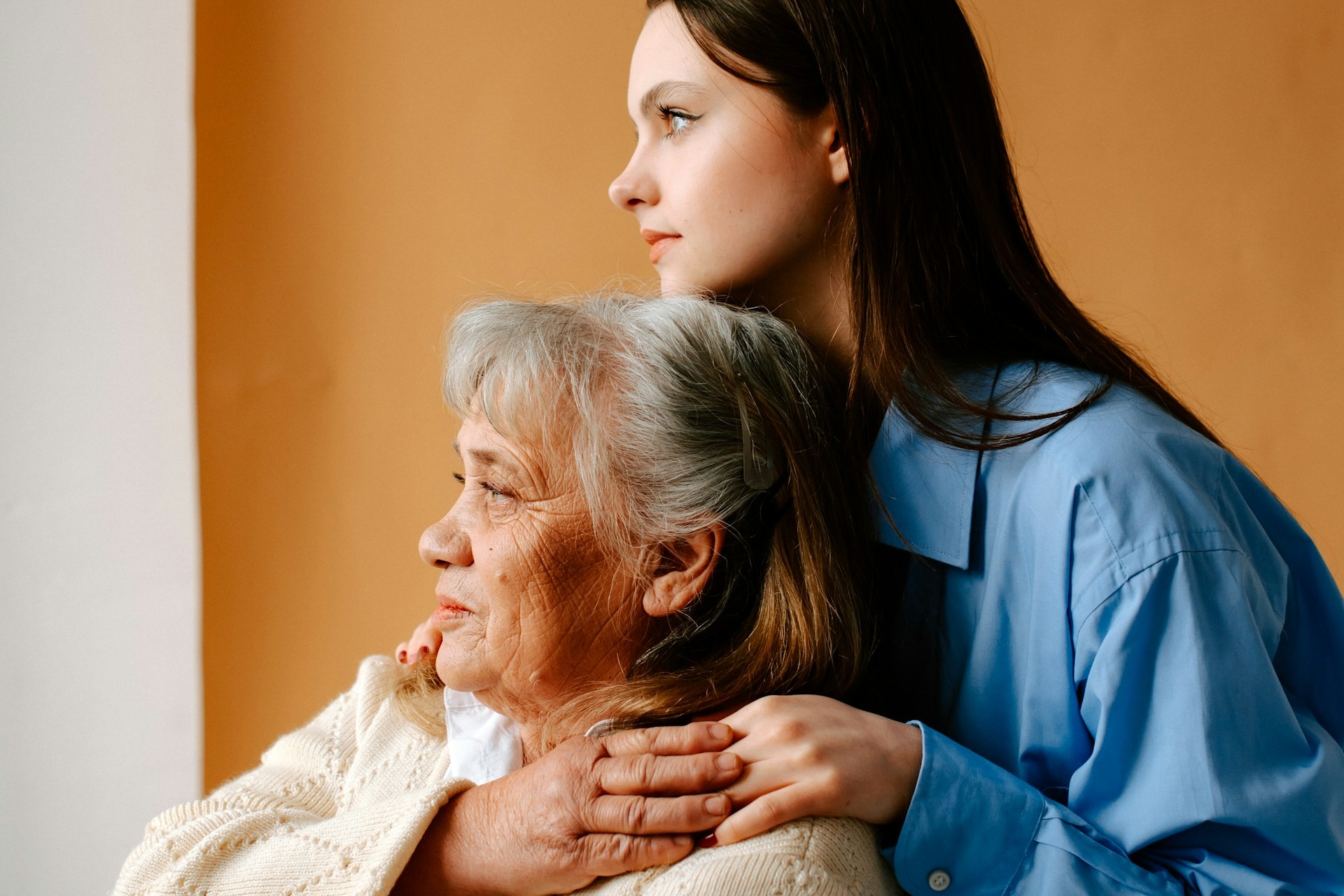 A young woman embraces an elderly woman from behind in a warm moment of connection, highlighting the support, compassion, and family bonds between generations.