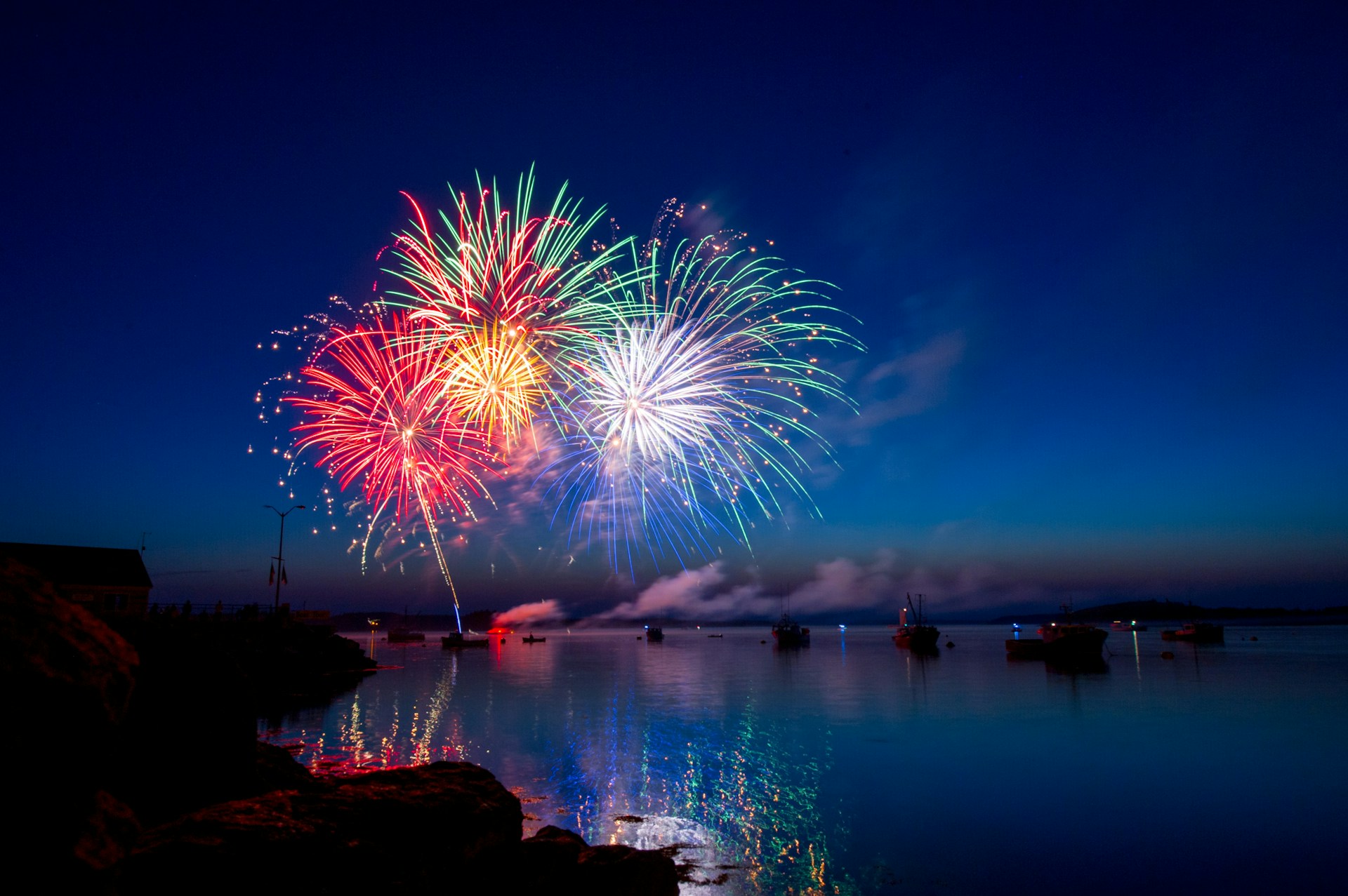 Colorful fireworks bursting over a calm body of water at night, with reflections shimmering on the surface and boats silhouetted against the illuminated sky.