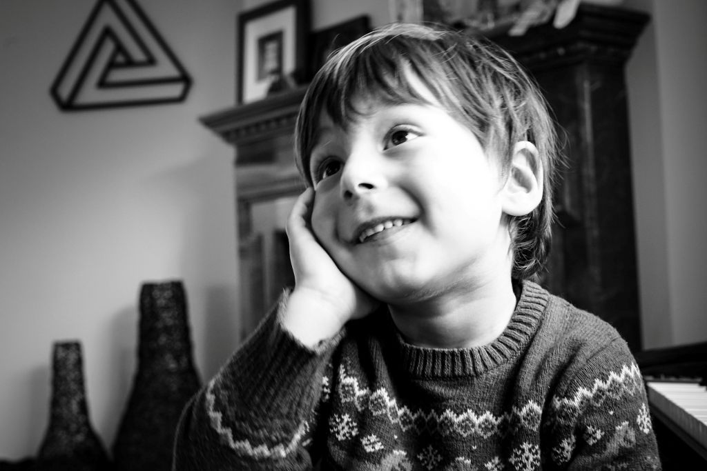 Smiling child in a patterned sweater rests their face on one hand while gazing upward with a curious, joyful expression in a cozy home.